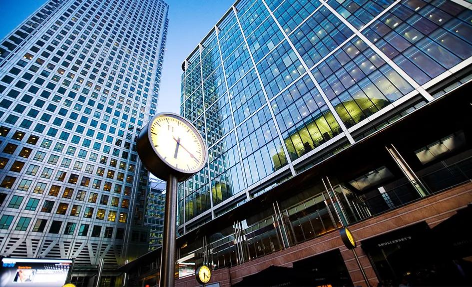 Modern glass skyscrapers and office buildings in Canary Wharf, City of London, with a large outdoor clock in the foreground.