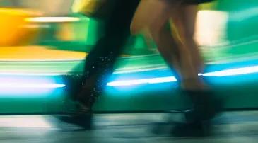 Blurred legs of a person walking past a brightly lit background with green, yellow, and blue light streaks