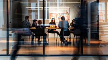Office meeting scene viewed through a glass wall. Five people are seated around a table, actively discussing or working. A laptop is open on the table, with papers and notebooks scattered. Blurred figures walk past outside the room, indicating motion. 