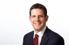 Mark Francis is a white man with short brown hair. He is wearing frameless glasses, a dark suit and a dark red tie. 